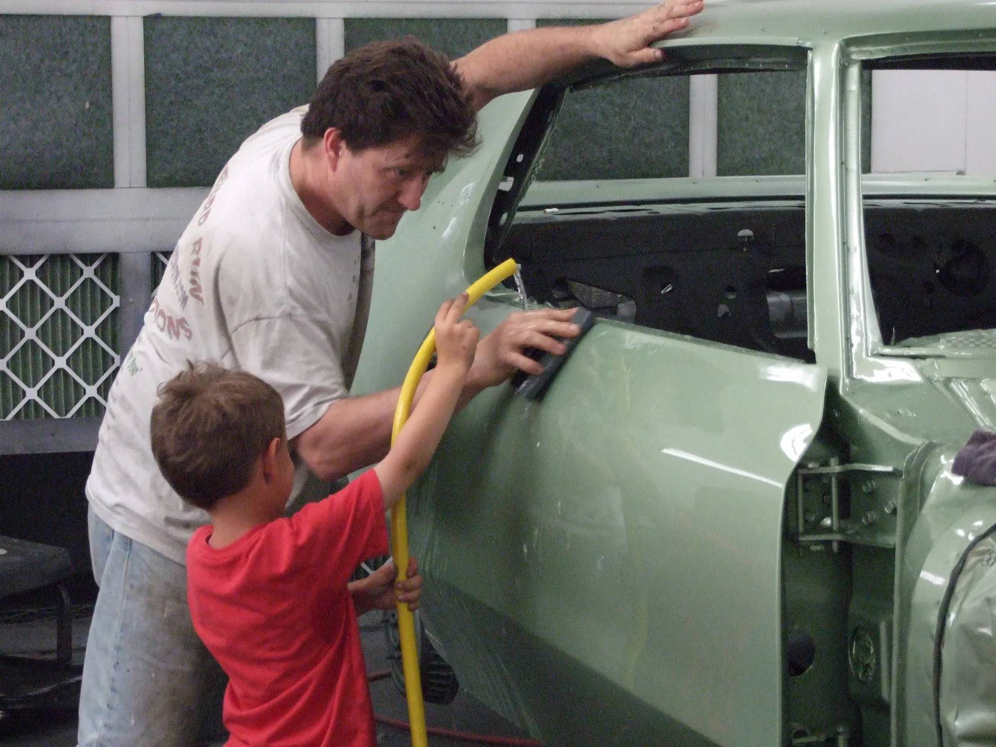 Mark Seybold and his young son wet-sanding the side of a classic car together on the shop floor