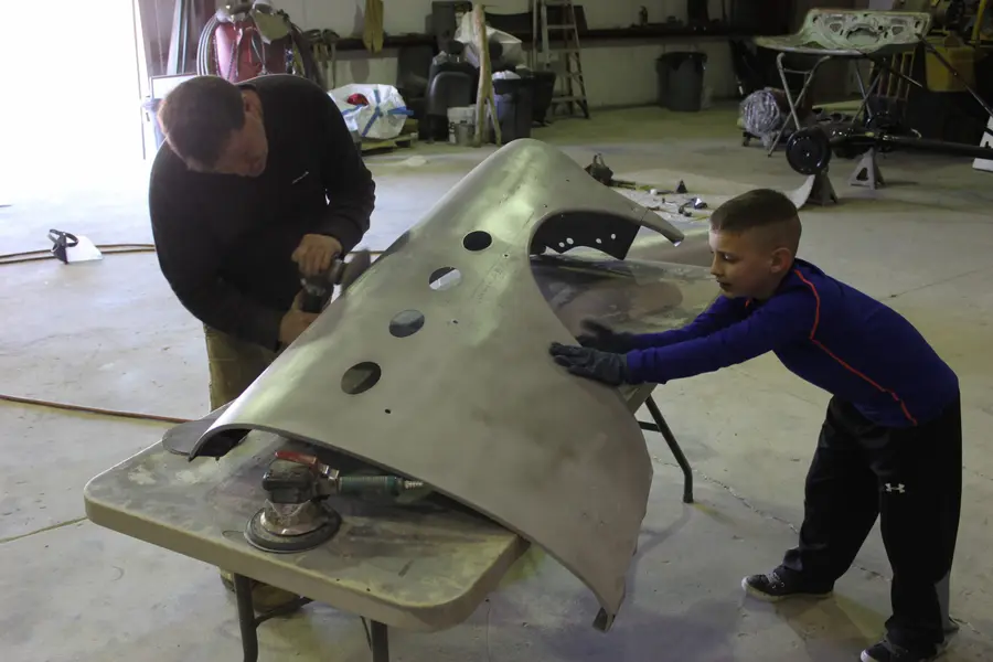 A craftsman and a child steadying a bare fender panel together across a work table